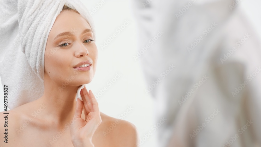 Gorgeous slim Caucasian young woman with towel on her head rubs her face with cotton pad smiling in front of the mirror in the bathroom | Skin moisturizing concept Gorgeous slim Caucasian young woman with towel on her head rubs her face with cotton pad smiling in front of the mirror in the bathroom | Skin moisturizing concept