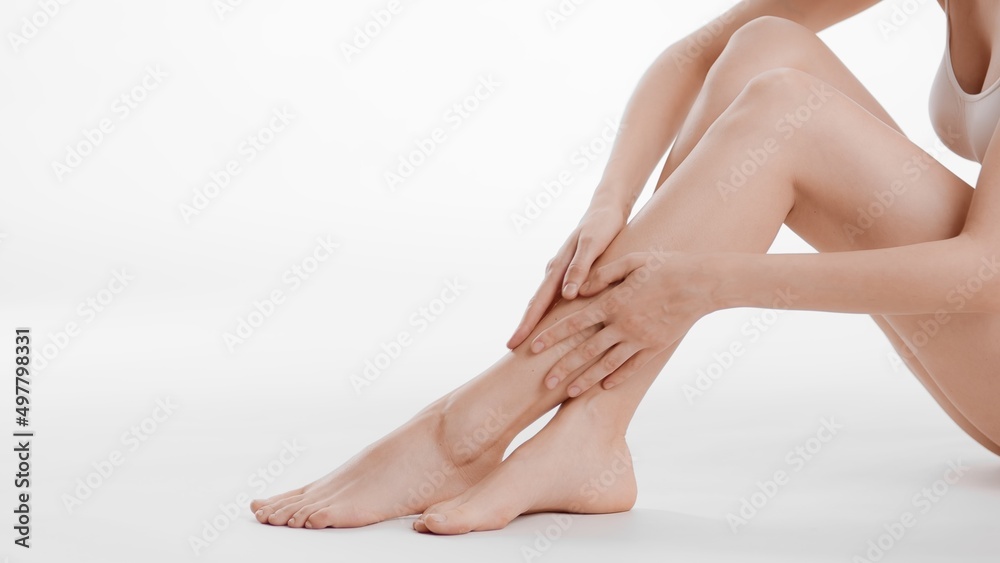 Medium shot of smooth female legs. Slim woman touches her crossed legs sitting on the floor on white background | Body care concept