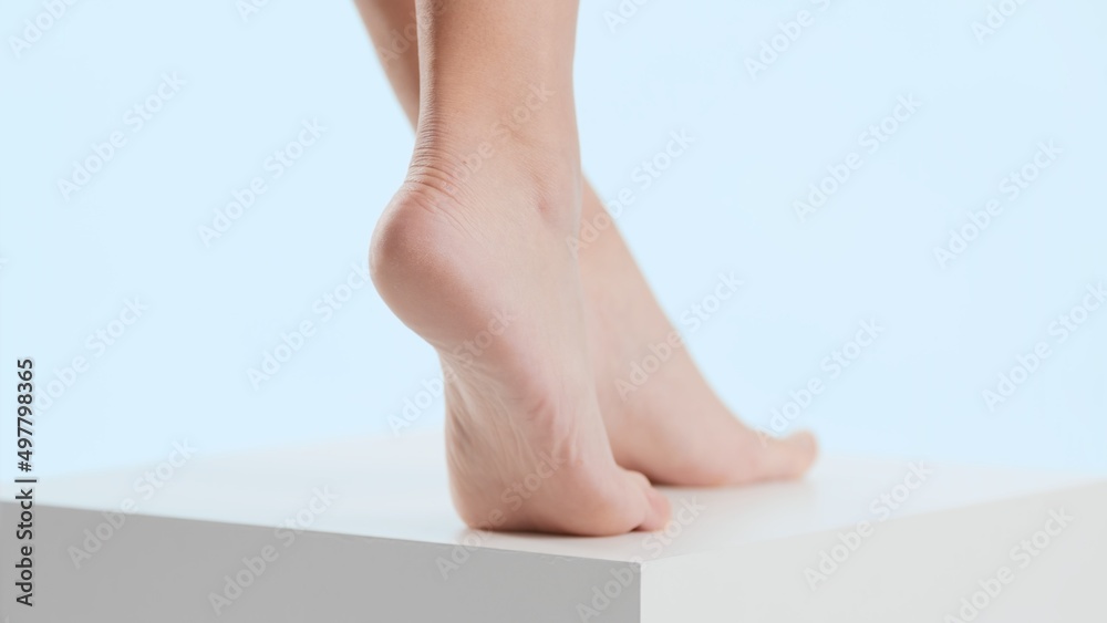 Close-up body part shot of female feet standing on cube platform against pale blue background | Anti cracked and smooth heel concept Close-up body part shot of female feet standing on cube platform against pale blue background | Anti cracked and smooth heel concept