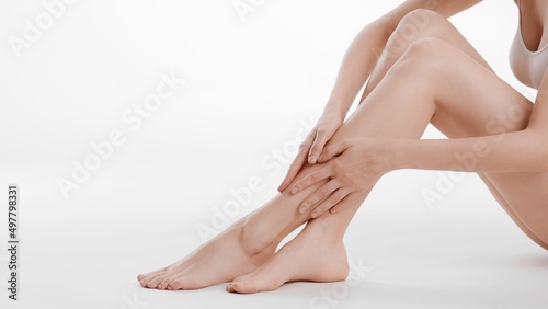 Medium shot of smooth female legs. Slim woman touches her crossed legs sitting on the floor on white background   Body care concept © pavel_dp