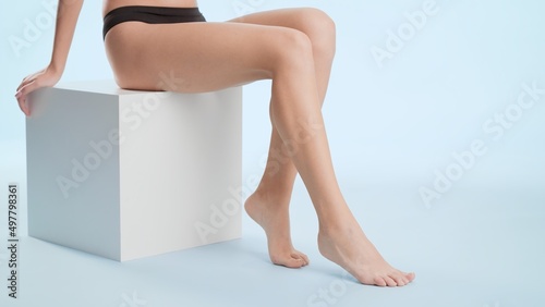 Flawless female legs. Young slim woman in black underwear sitting on the white cube platform on pale blue background | Leg care and unwanted hair removal concept