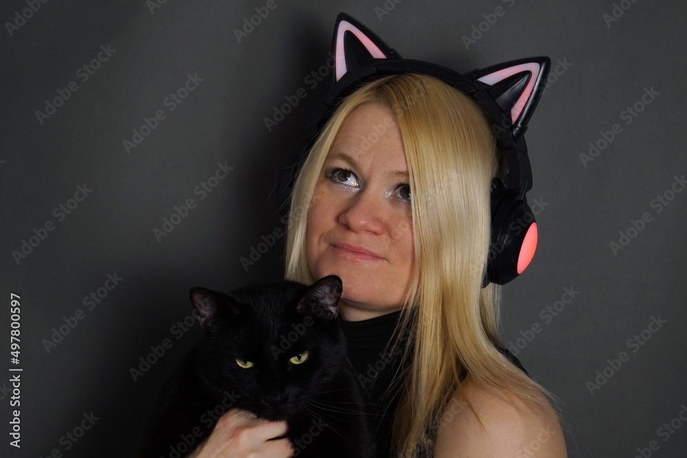 Dark night. Woman wearing headphones in the shape of cat ears with a black cat in her arms.