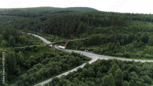 Aerial view of a countryside landscape with the highway and green forest. Scene. A truck and a passenger car driving on highway through scenic rolling green hills with trees and vegetation.