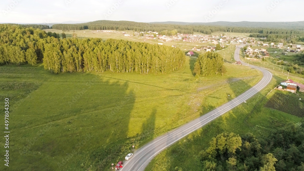 Beautiful summer landscape from a bird's-eye view. Scene.A long road with driving cars and a huge beautiful green forest with tall trees and behind which a village with small houses and a blue clear