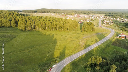 Beautiful summer landscape from a bird s-eye view. Scene.A long road with driving cars and a huge beautiful green forest with tall trees and behind which a village with small houses and a blue clear