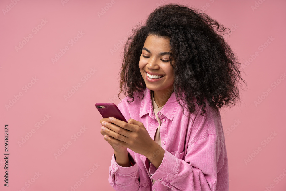 Beautiful smiling African American woman using smartphone playing mobile game isolated on pink background. Happy stylish female holding mobile phone shopping online with sale, copy space Beautiful smiling African American woman using smartphone playing mobile game isolated on pink background. Happy stylish female holding mobile phone shopping online with sale, copy space
