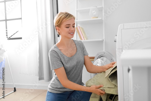 Young woman putting green jacket into washing machine in bathroom