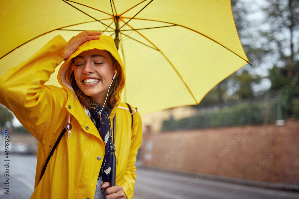 Close-up of a young cheerful woman with a yellow raincoat and umbrella who is in a good mood while walking the city on a rainy day and posing for a photo. Walk, rain, city - obrazy, fototapety, plakaty 