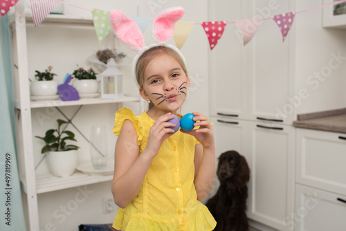a happy girl with long blonde hair in the image of an Easter bunny holds decorated eggs in her hands  looking into the camera makes a kiss  happy Easter at home.