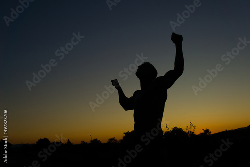 Silhouette of muscular man posing shows his muscles against the sky at sunset. 