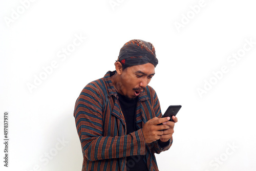 Surprised Asian man in javanese traditional costume standing while holding a cellular phone.
