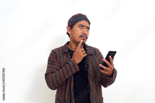 Thoughtful Asian man standing while holding a cellular phone. Isolated on white background © SetianingDyah