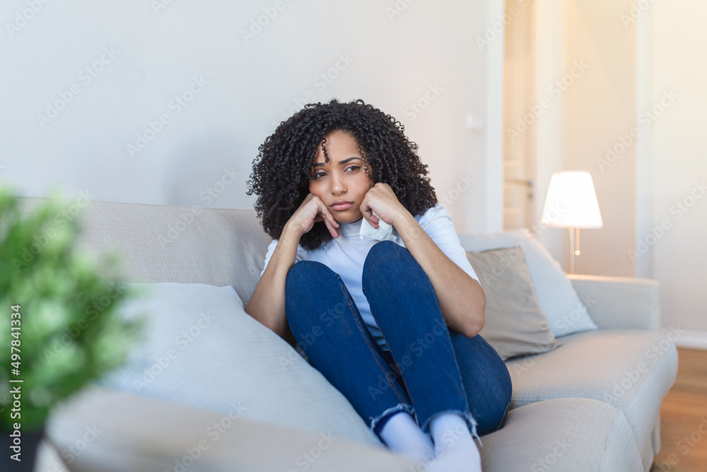 Unhappy african American woman on sofa crying. Lonely sad woman deep in thoughts sitting daydreaming or waiting for someone in the living room with a serious expression, sitting on couch Unhappy african American woman on sofa crying. Lonely sad woman deep in thoughts sitting daydreaming or waiting for someone in the living room with a serious expression, sitting on couch