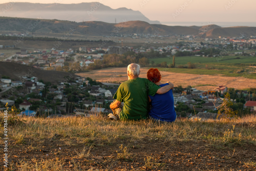 An elderly couple sits on a mountain with their backs with a beautiful view of the mountains and the sea in the distance.