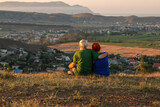 An elderly couple sits on a mountain with their backs with a beautiful view of the mountains and the sea in the distance.