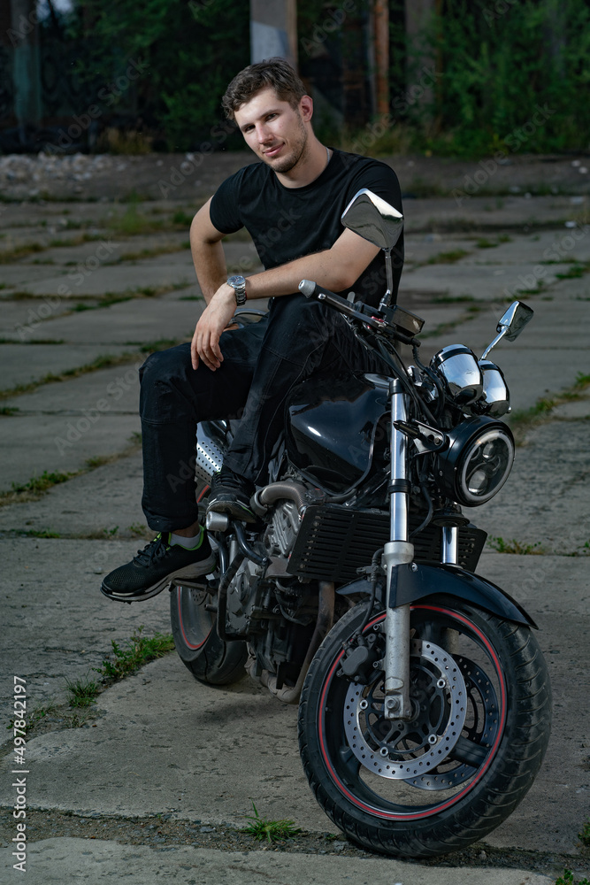 A young motorcyclist in a black T-shirt sits on a classic motorcycle parked in an abandoned area