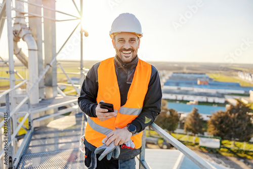 Obraz na płótnie An industry worker holding phone on metal construction and smiling at the camera