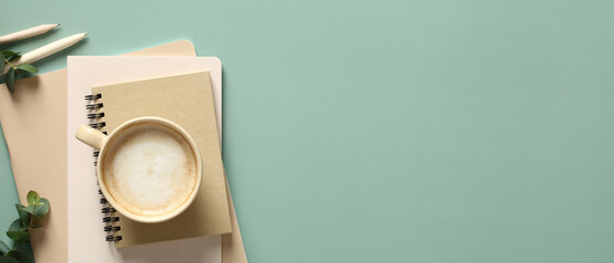 Banner with cup of coffee, stack of paper notebooks and eucalyptus leaves on green background. Home office desk table, nordic style.