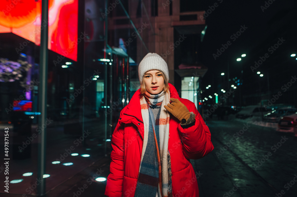 Night portrait of a young woman in warm clothes on the streets of the metropolis while walking, looking at the camera.