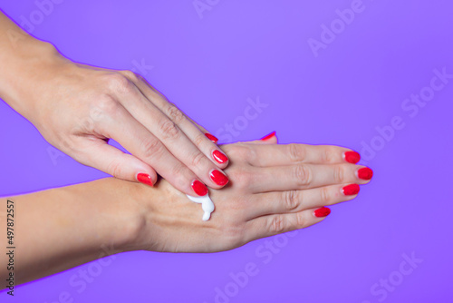 Hands of young woman with red manicure apply cream to skin on purple background
