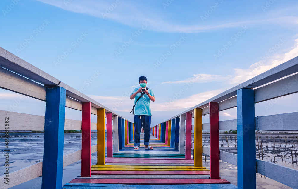 Perspective view of Asian young man in protective mask using mini digital camera to taking video on the old multicolored wooden bridge at sea viewpoint against blue sky Perspective view of Asian young man in protective mask using mini digital camera to taking video on the old multicolored wooden bridge at sea viewpoint against blue sky