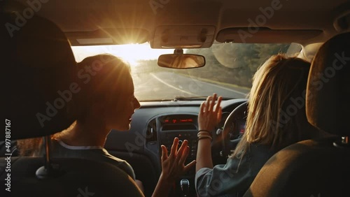 Rear view of cheerful young girlfriends singing their favourite song and dancing while driving at sunset. Two beautiful women travelling and having good time together . Concept of friendship photo