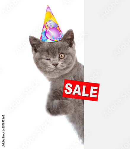 Winking kitten wearing sunglasses and birthday cap looks from behind empty white banner and holds sales symbol. isolated on white background