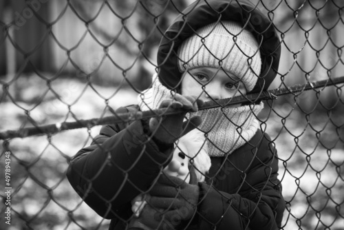 Little refugee girl with a toy behind a metal fence. Social problem of refugees and internally displaced persons. Russia's war against the Ukrainian people photo