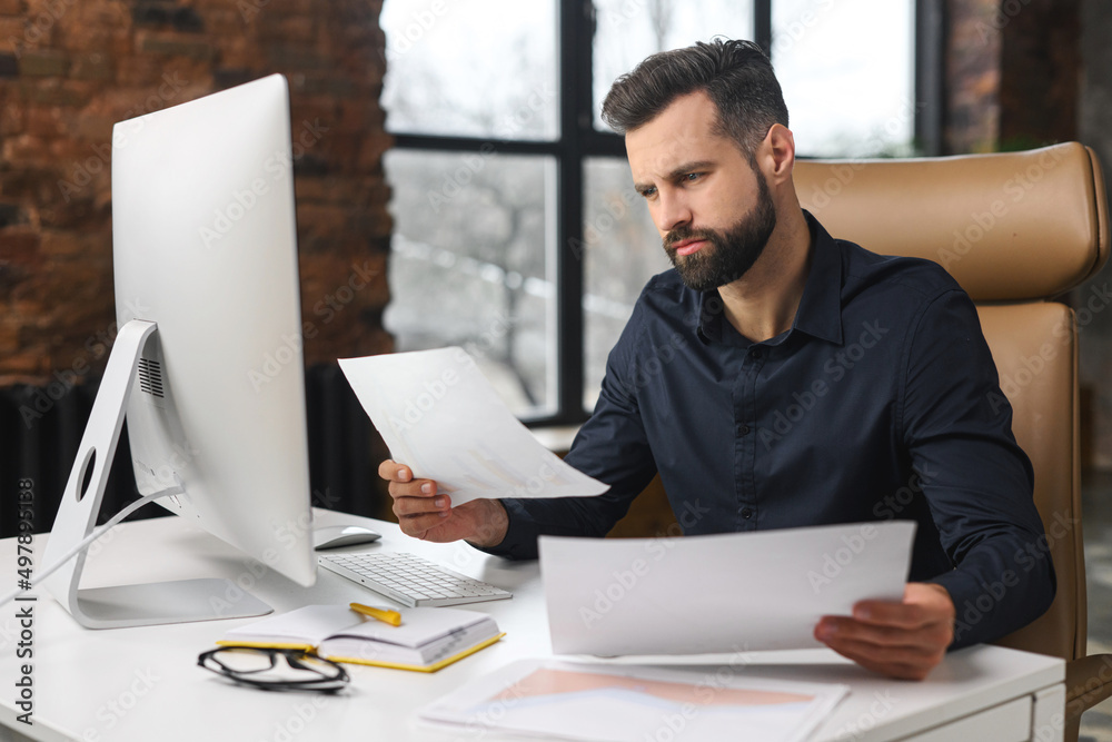 Concentrated caucasian guy is doing paperwork. Man looking through paper sitting at the desk with a computer in office. Man checking report concept Concentrated caucasian guy is doing paperwork. Man looking through paper sitting at the desk with a computer in office. Man checking report concept