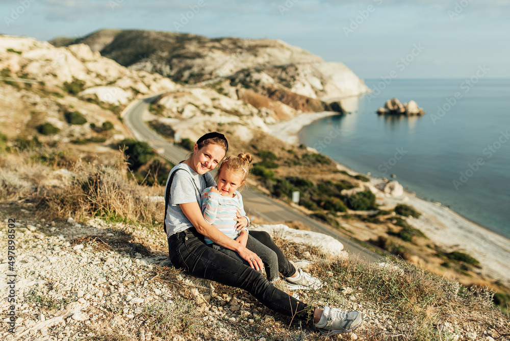 family, little girl sitting in mom's arms on the background of rocky sea shore, sunny day, positive emotions