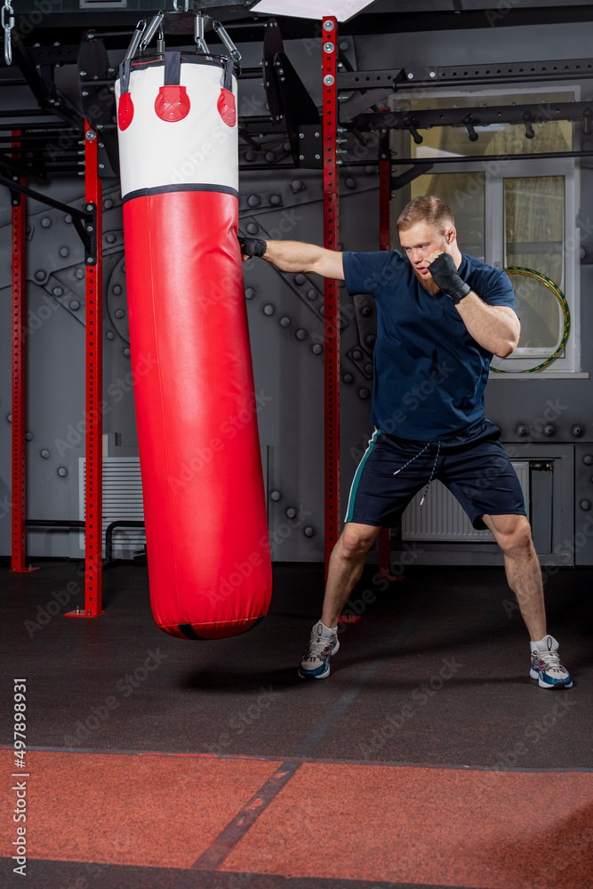 Young strong bearded man, mixed martial arts fighter and trainer, practices punches with his right hand on red punching bag in sports hall.
