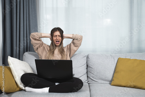 Young student or business woman sitting at home covering her ears with hands because she cant study or work on her laptop computer from loud noise coming from apartment above. photo