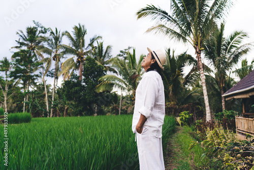 Side view of happy Balinese farmer in casual hat smiling during daytime in Thailand, cheerful adult man in white apparel visiting countryside with rice fields - natural ecology for cultivation