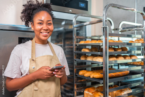 Fotografiet Close up of joyful African American young woman worker in apron stands in bakery shop, speking on smartphone and using tablet device