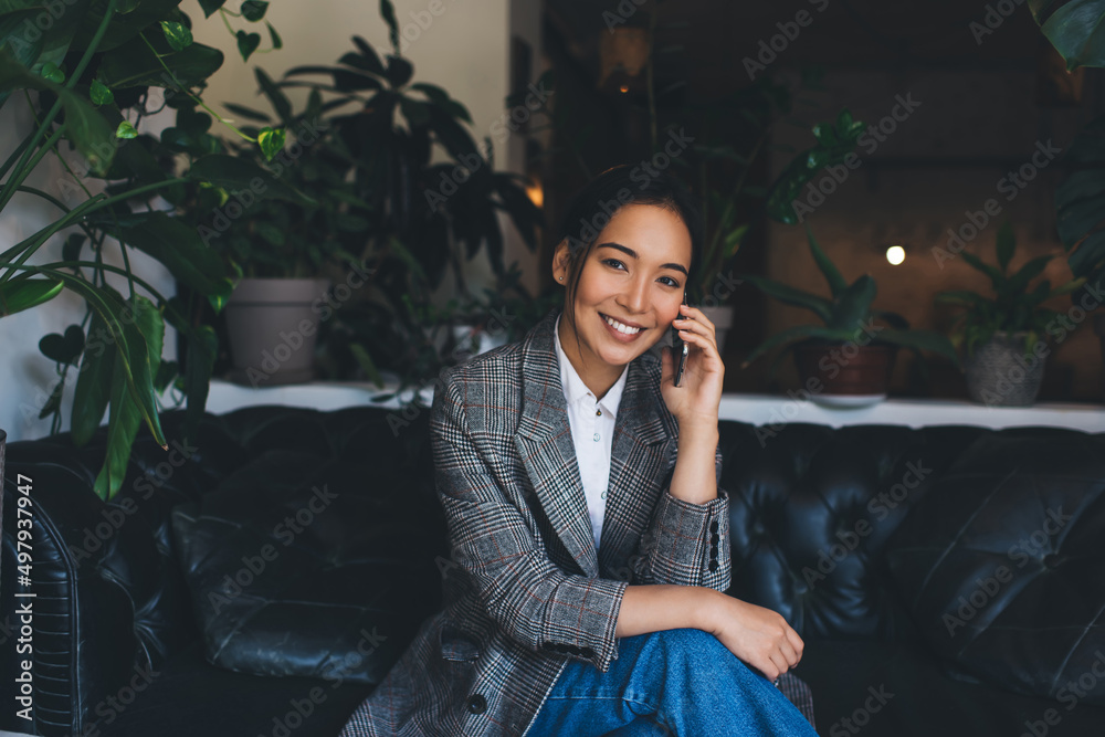 Portrait of happy Asian woman in jacket smiling at camera while calling to friend for talking indoors, successful female enjoying mobile communication using smartphone application and wireless Portrait of happy Asian woman in jacket smiling at camera while calling to friend for talking indoors, successful female enjoying mobile communication using smartphone application and wireless