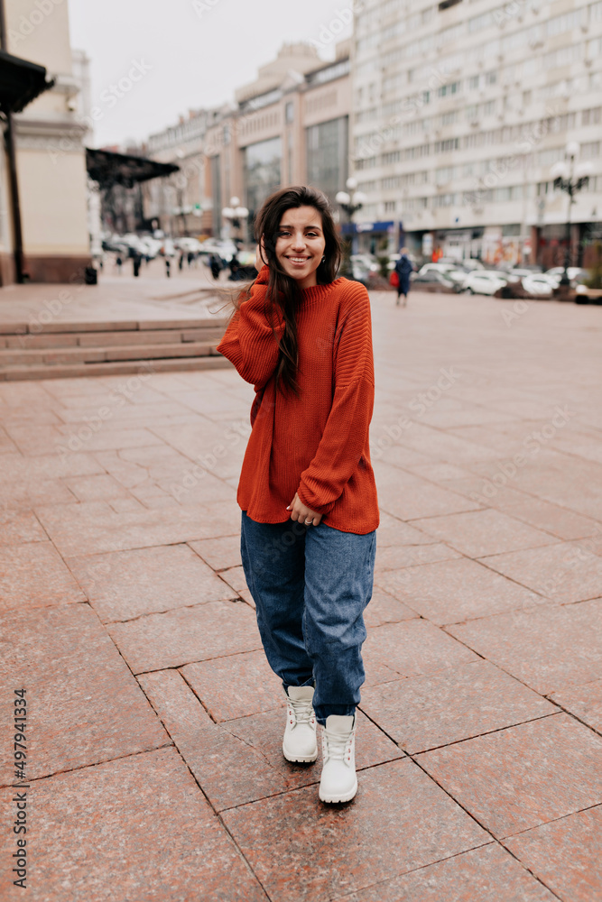 Full-lenght portrait of charming pretty girl with long dark hair and wonderful smile is wearing orange knitted sweater and jeans is walking on the street in warm day