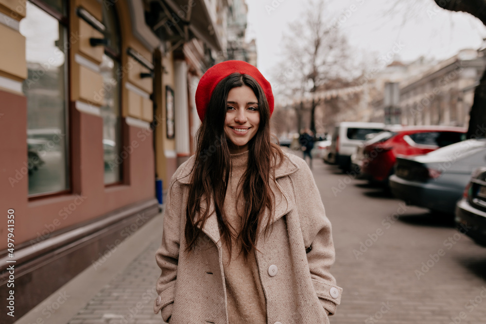 Fototapeta premium Charming pretty european woman with dark long hair and wonderful smile is wearing red beret and beige coat is walking down the city centre 
