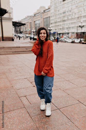 Full-lenght portrait of charming pretty girl with long dark hair and wonderful smile is wearing orange knitted sweater and jeans is walking on the street in warm day © PhotoBook