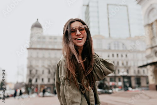 Cheerful brunette woman with long flying hair in brown glasses and green jacket smiles sincerely outdoors. Happy lady laughs and walks in great mood outside. © PhotoBook
