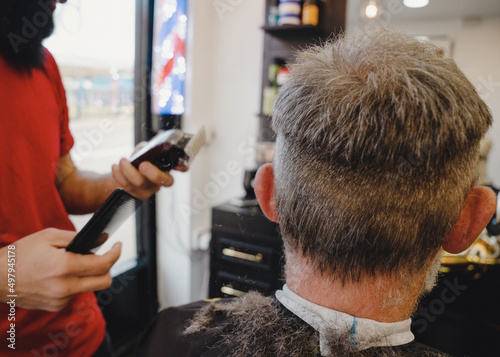 Barber trimming and cutting bearded man with shaving machine in barbershop. Hairstyling process.