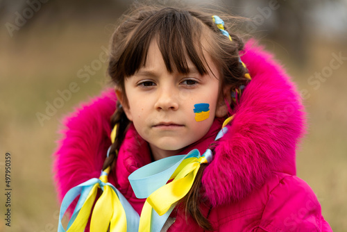 A little Ukrainian refugee girl with a Ukrainian flag painted on her cheek is sad about her lost home. Stop the war in Ukraine. Help save the Ukrainian nation