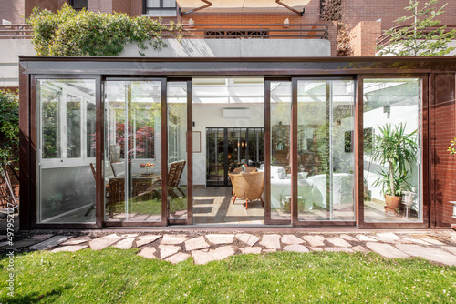 Glass and brown aluminum covered terrace at the bottom of a residential building with sliding doors, decorative trees and a paver path and decorative plants, rattan and wooden furniture inside