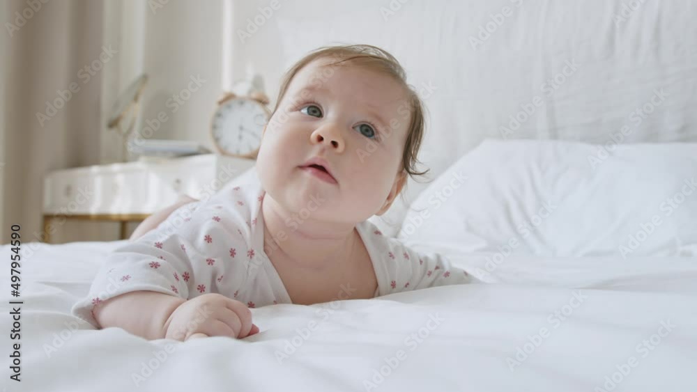 Cute sweet baby newborn with blue eyes lying on white bed sheet expressive looks around with interest and moving her lips. Third month of life closeup face portrait. Baby is lying on his tummy Cute sweet baby newborn with blue eyes lying on white bed sheet expressive looks around with interest and moving her lips. Third month of life closeup face portrait. Baby is lying on his tummy