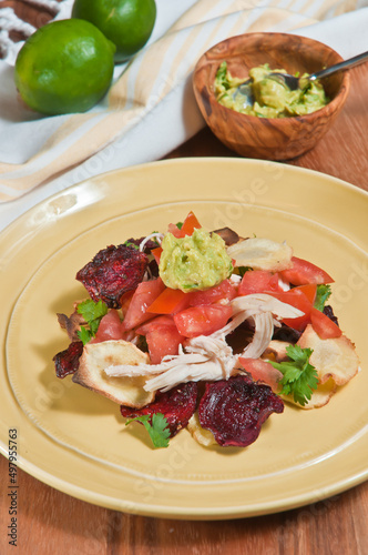 top view, close distance of a yellow , ceramic plate filled with sliced beets and parsnip, nacho chips with chopped tomatoes, lettuce, shredded chicken and wood bowl of guacamole