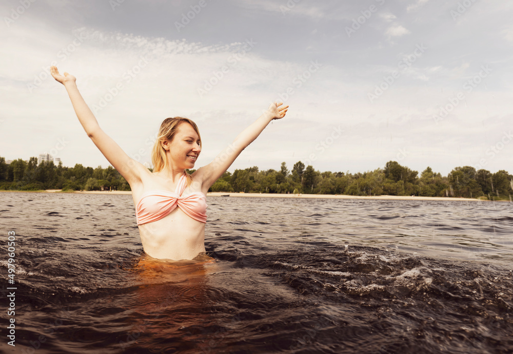 A young blonde woman joyfully raises her hands up while standing in dark water. Woman swimming in summer A young blonde woman joyfully raises her hands up while standing in dark water. Woman swimming in summer