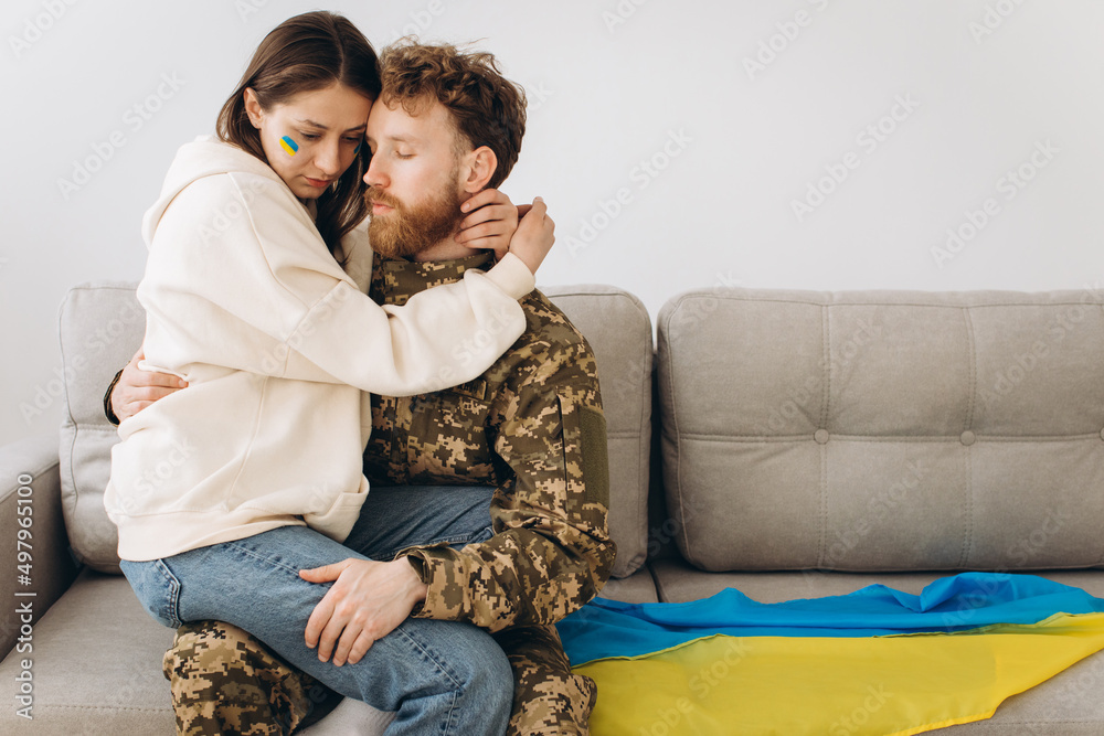 Ukrainian couple, military man in uniform with his girlfriend on the couch at home on a background of yellow and blue flag Ukrainian couple, military man in uniform with his girlfriend on the couch at home on a background of yellow and blue flag