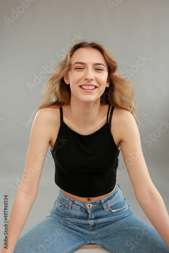 Beautiful girl model appearance in light jeans and black T-shirt on a gray studio background. Girl model sitting on a white chair. Beautiful blonde shows poses for a photo shoot © Anhelina Tyshkovets