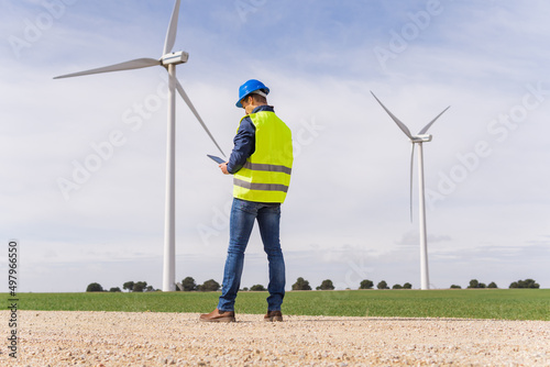 Unrecognizable worker from the renewable energy sector, from the back looking at his digital tablet in a field of wind turbines. Increase in energy prices in the market and alternative energies.