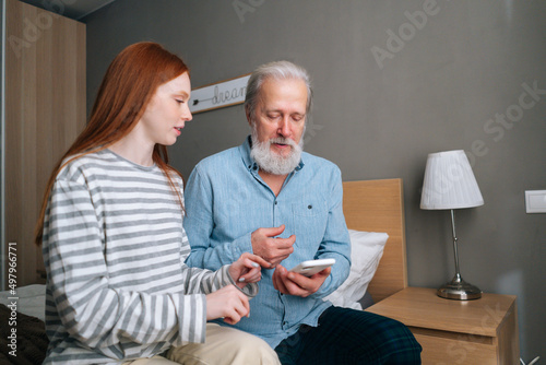 Caucasian bearded senior grandfather learning to using mobile phone under guidance of cute young granddaughter sitting on bed at home. Portrait of female teaching grandpa using smartphone. © dikushin