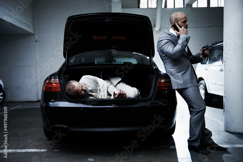 Hostage to the mob. A frightened businessman lying in the trunk of a car while his armed captor talks on a cellphone.